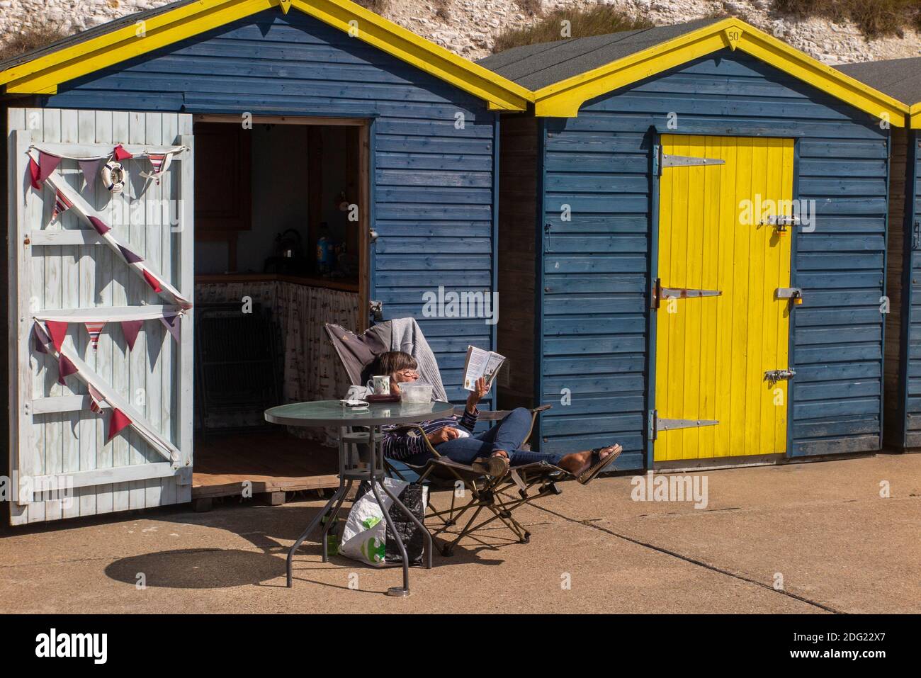 Genießen Sie die Sonne und lesen Sie in einem Liegestuhl vor einer Strandhütte an der Küste von Kent in der Nähe von Margate, Thanet, England Stockfoto