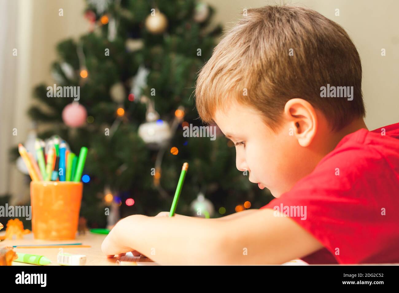 7 Jahre alter Junge, der am Schreibtisch sitzt und stumpf wird. Kind schreibt Notizen in Notizbuch, Nahaufnahme, Seitenansicht. Geschmückter Weihnachtsbaum im Hintergrund Stockfoto