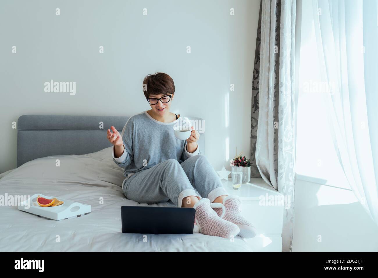 Emotionale Frau in Brillen, lässige Hauskleidung mit Video-Chat auf ihrem Laptop sitzen auf dem Bett und Kaffee trinken. Telearbeit von zu Hause aus. Zeit um Stockfoto