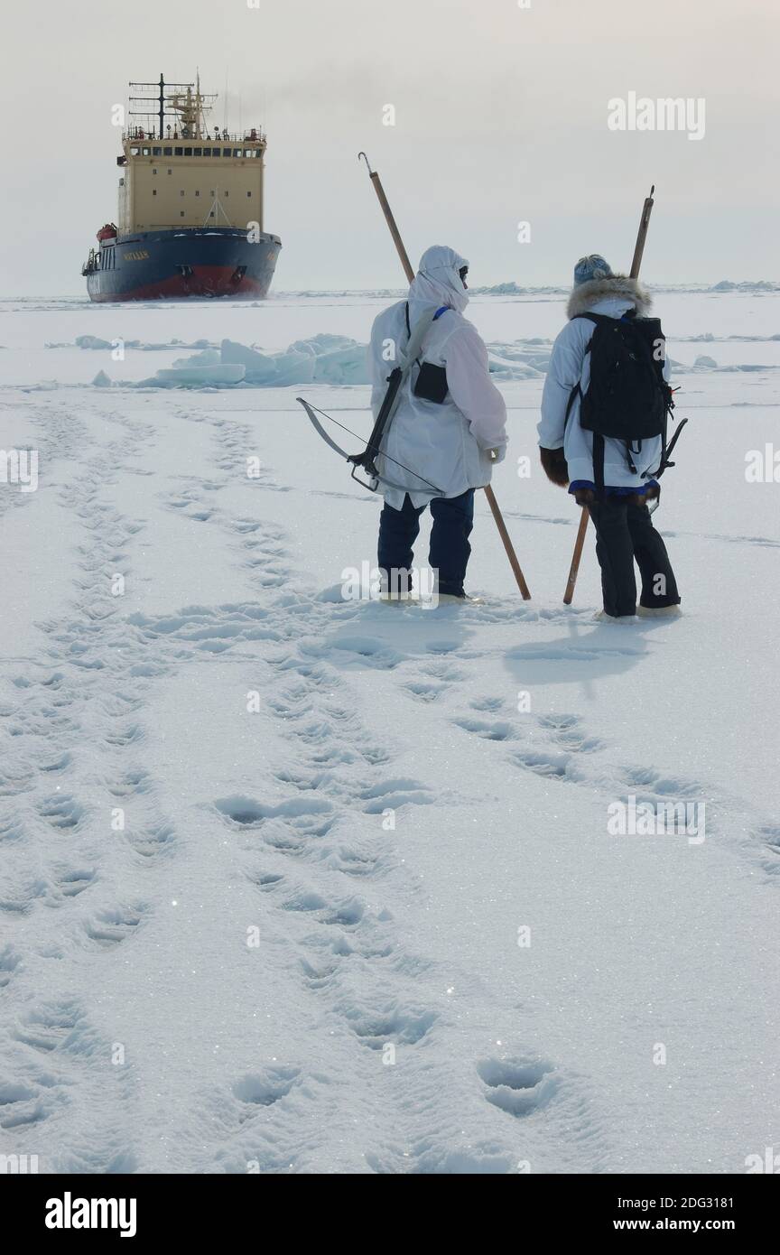 Arctic native Ice Test Stick. Zwei Menschen auf dem Meereis in der nördlichen Beringsee halten in der Arktis einheimische Eisteststäbe, die als "unghaqs" bezeichnet werden. Die oben auf den Stöcken sichtbaren Haken dienen zum Auffinden von Dingen, wie zum Beispiel das Greifen der Kleidung einer Person, wenn sie ins Wasser fallen, und das andere Ende wird zum Stocken des Eises verwendet. Im Hintergrund der russische Eisbrecher Magadan. Unghaqs sind ein hoch geschätztes Sicherheitsinstrument für Alasker, die in Küstengemeinden leben und sich im Winter auf das Eis wagen, um dort zu jagen. Stockfoto