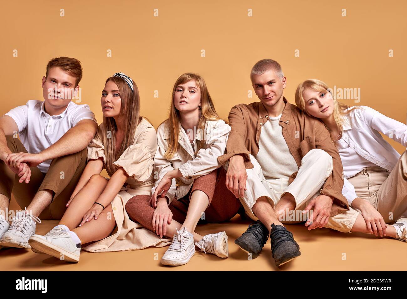 kaukasische junge Menschen sitzen zusammen auf dem Boden isoliert auf braun, tragen die gleichen stilvollen Outfits, Blick auf Kamera Stockfoto