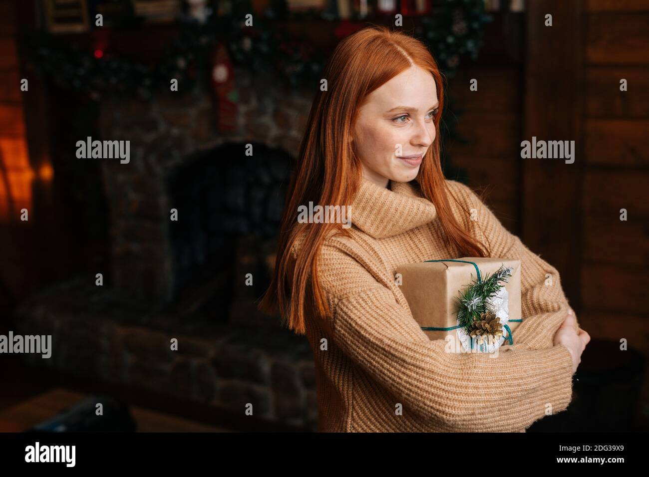 Lächelnd rothaarige junge Frau hält schöne Weihnachts-Geschenkbox, während sie im gemütlichen Wohnzimmer steht. Stockfoto