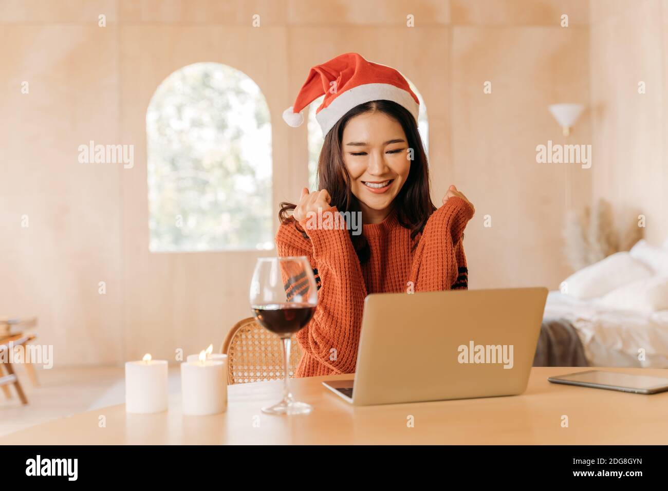 Junge Teenager asiatische Frau trägt Pullover mit hübschen Lächeln und santa Hut im Schlafzimmer. Glückliche und erfolgreiche Mädchen mit Laptop mit Kerzen und Wein auf dem Tisch. Winter Portrait Mode Lifestyle. Speicherplatz kopieren Stockfoto