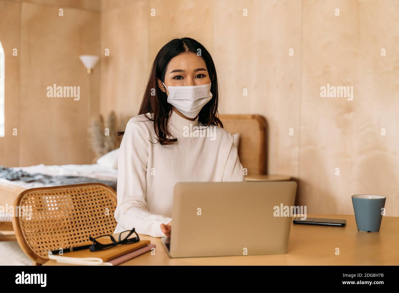 Junge Teenager asiatische Frau trägt Pullover mit Computer-Laptop arbeiten von zu Hause aus drinnen online. Attraktives Mädchen trägt eine Gesichtsmaske, um die Ausbreitung von Covid-19 - Coronavirus Pandemie zu verhindern. Speicherplatz kopieren Stockfoto