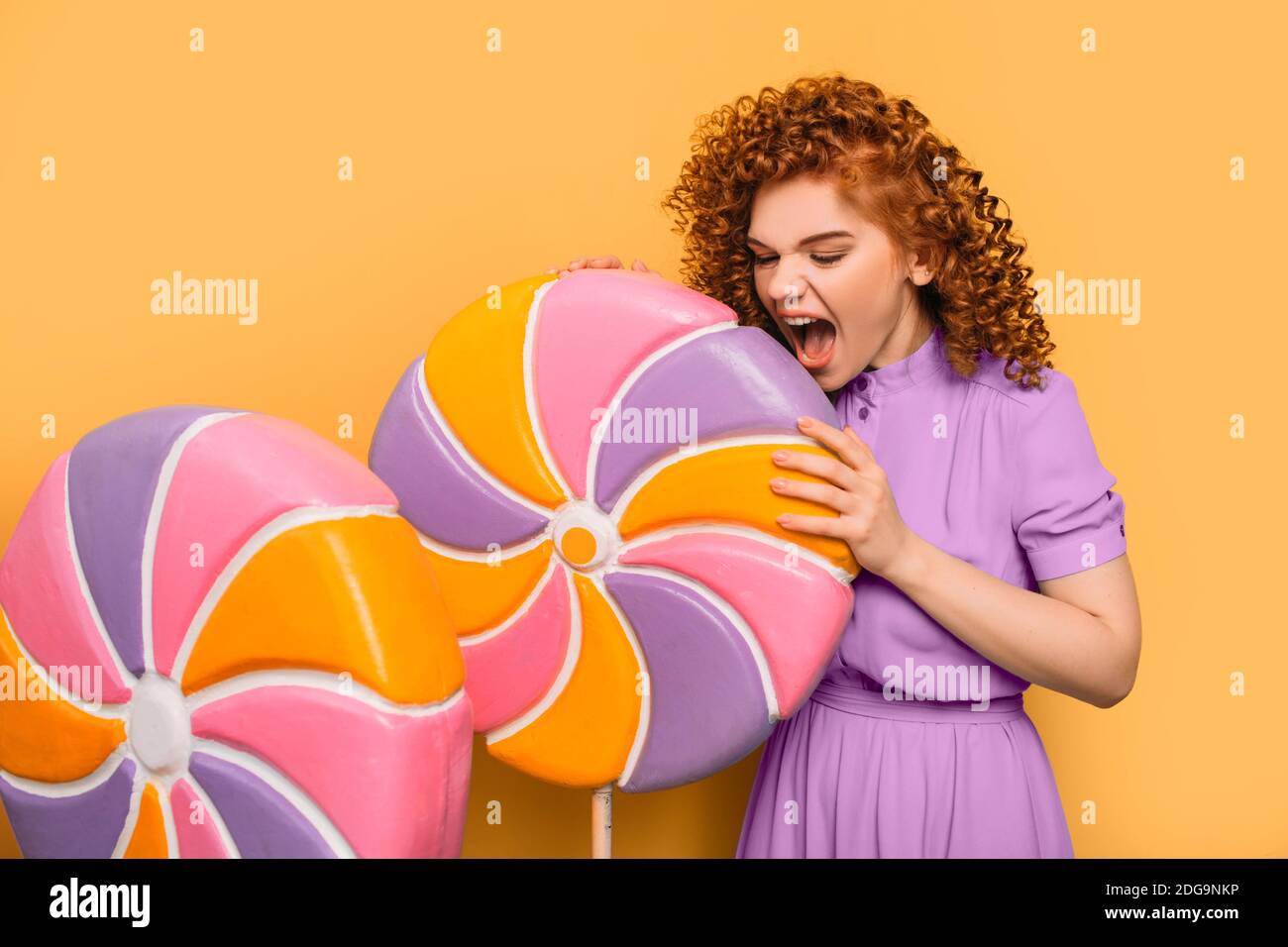 Lollipop, ich will dich essen. Lockige rothaarige positive Frau trägt ein violettes Kleid mit süßen großen Lutscher auf orangefarbenem Hintergrund Stockfoto