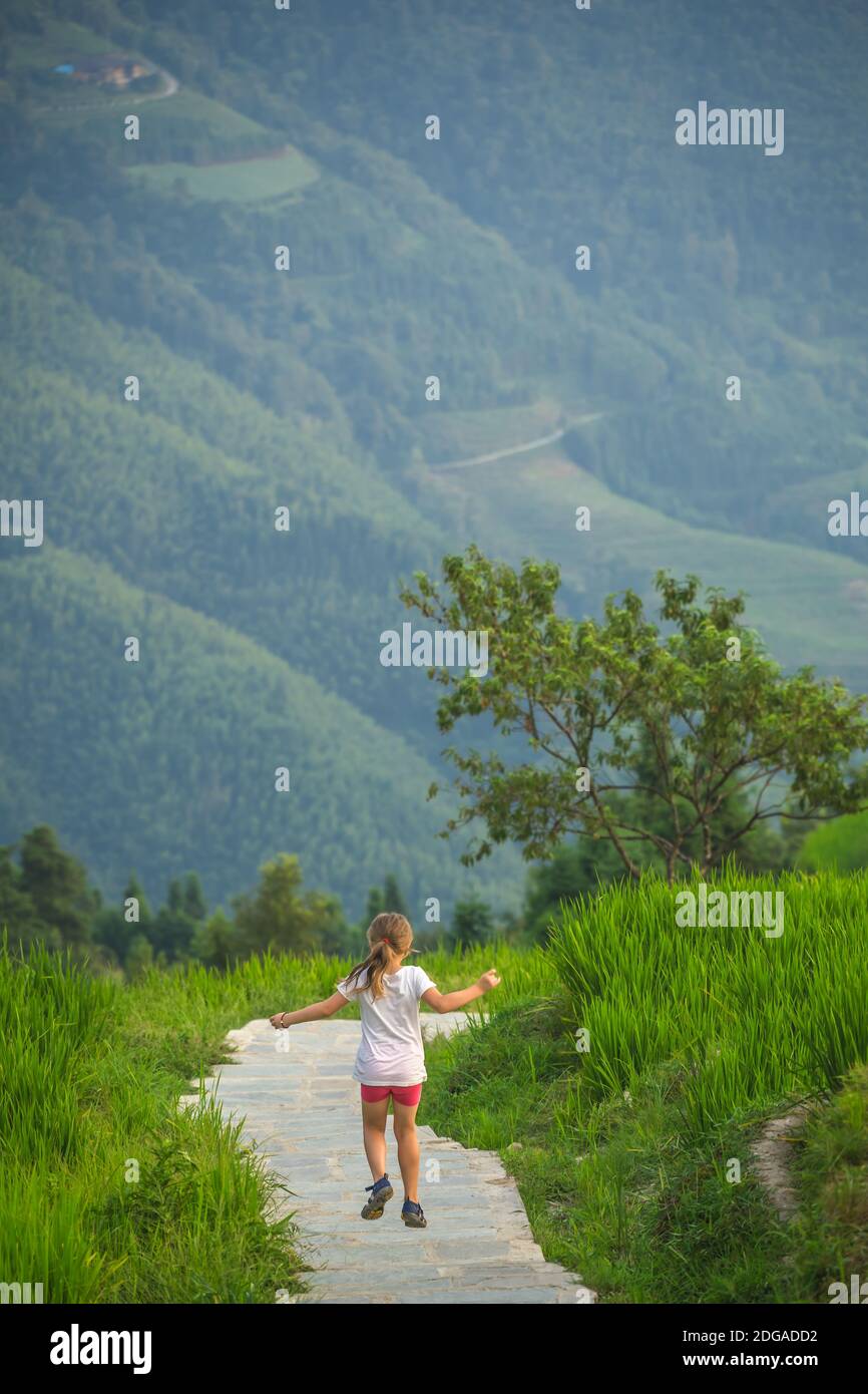 Niedliches junges Mädchen, das auf einem schmalen Pfad durch die kaskadierenden geschichteten Longji-Reisterrassen, Pingan-Dorf, nördliches Guilin, Guangx, springt und läuft Stockfoto