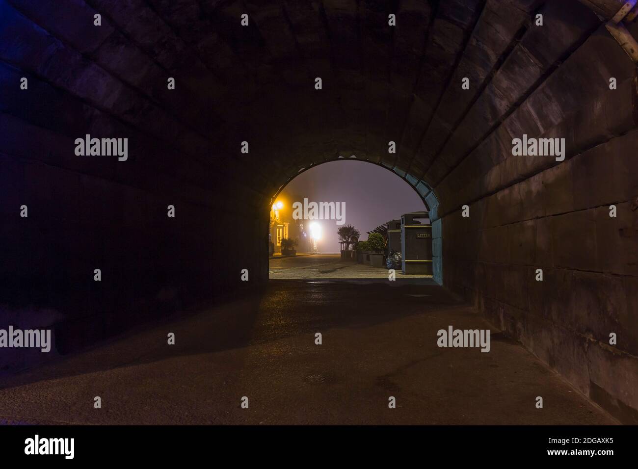 Stimmungsvolle, abendliche Straßenszene mit Blick durch einen alten Backsteintunnel bei Nacht. Blick auf die Straßenlaterne durch einen dunklen Steintunnel. Stockfoto