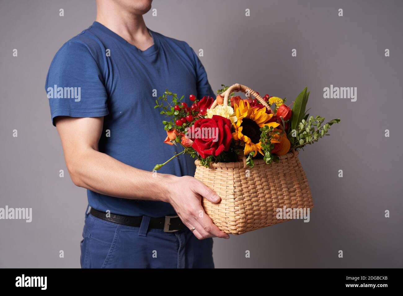 Ein weißer Mann in einem blauen T-Shirt hält einen Blumenkorb mit einem Kopierplatz auf einem grauen Hintergrund aus, Blumenlieferung Stockfoto
