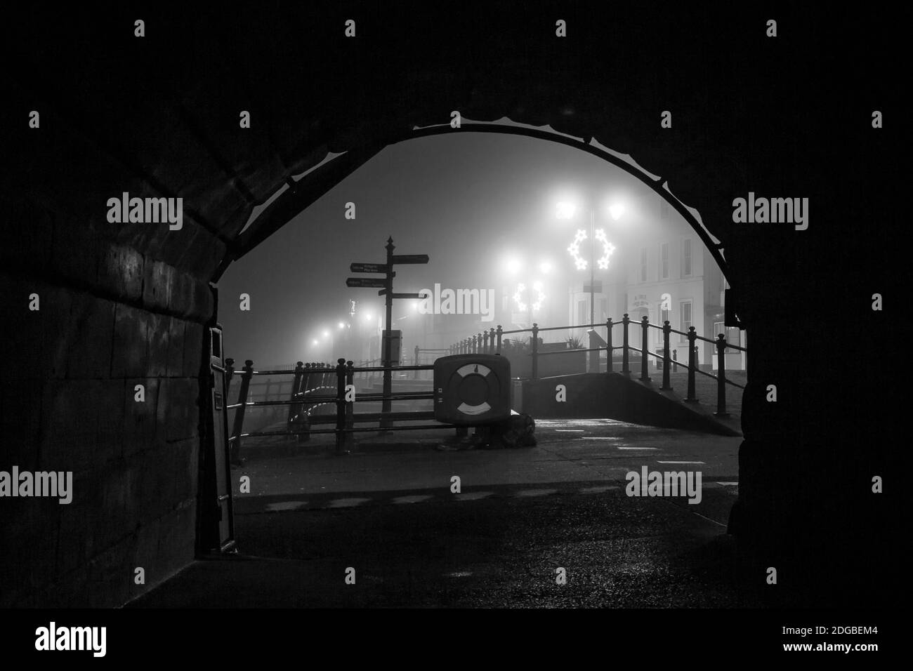 Monochrome, alte britische Straßenszene bei Nacht mit Straßenbeleuchtung, die durch einen Brückentunnel betrachtet wird. Stockfoto