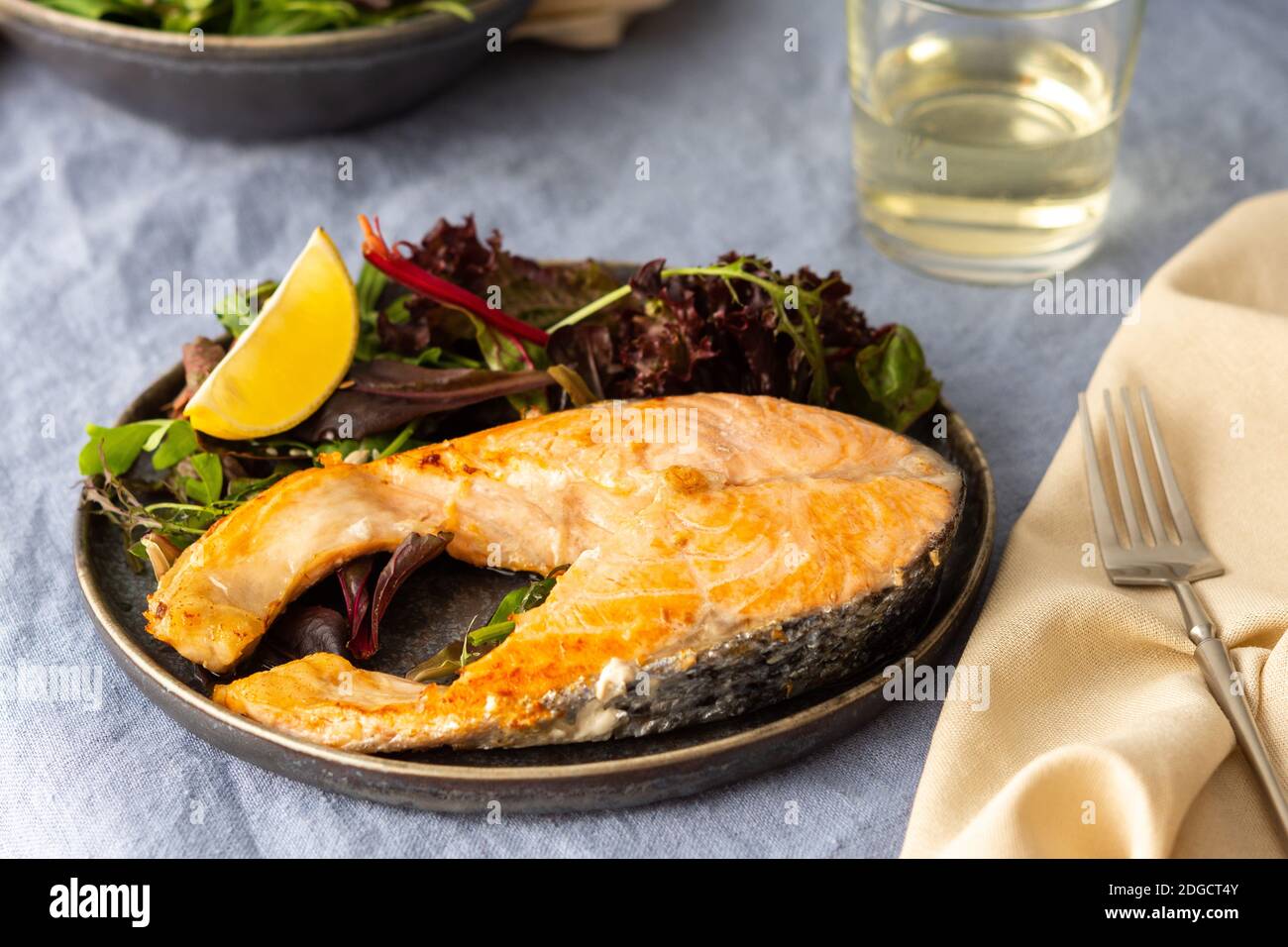 Gebratenes Lachssteak mit Salat auf einem Teller in Nahaufnahme, Weißwein im Glas, leckeres herzhaftes Abendessen oder Mittagessen Stockfoto