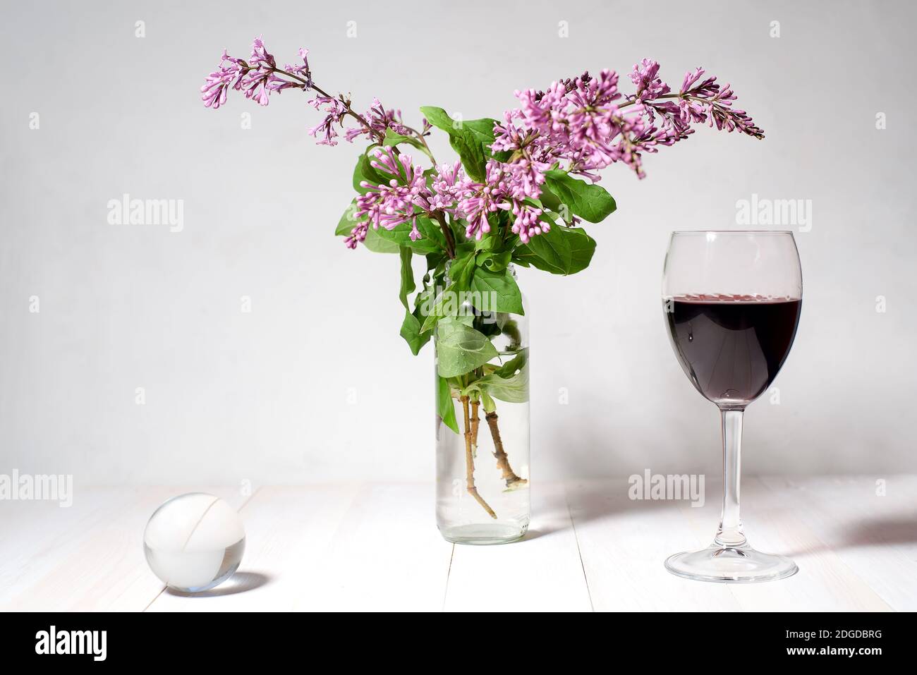 Stillleben aus einem Blumenstrauß Flieder in einer Vase, einem Glas Wein und einer transparenten Kugel auf einem weißen Rücken Stockfoto