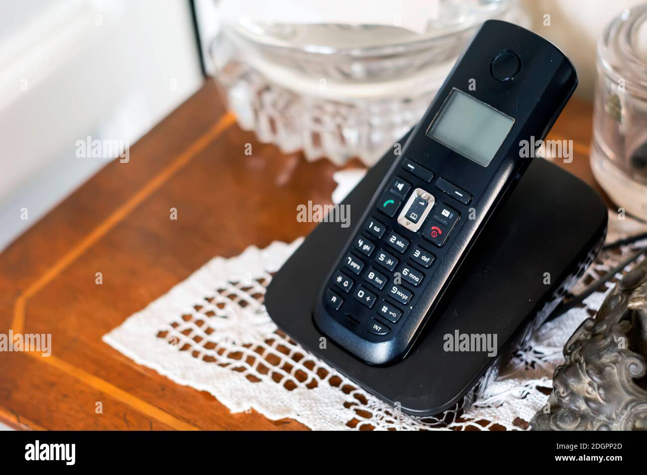 Ein schwarzes schnurloses Telefon aus Kunststoff, das auf seiner Wiege in einem klassisch eingerichteten Haus mit Antiquitäten und weißen Häkeln aufgeladen wird. Veraltete Technologie Stockfoto