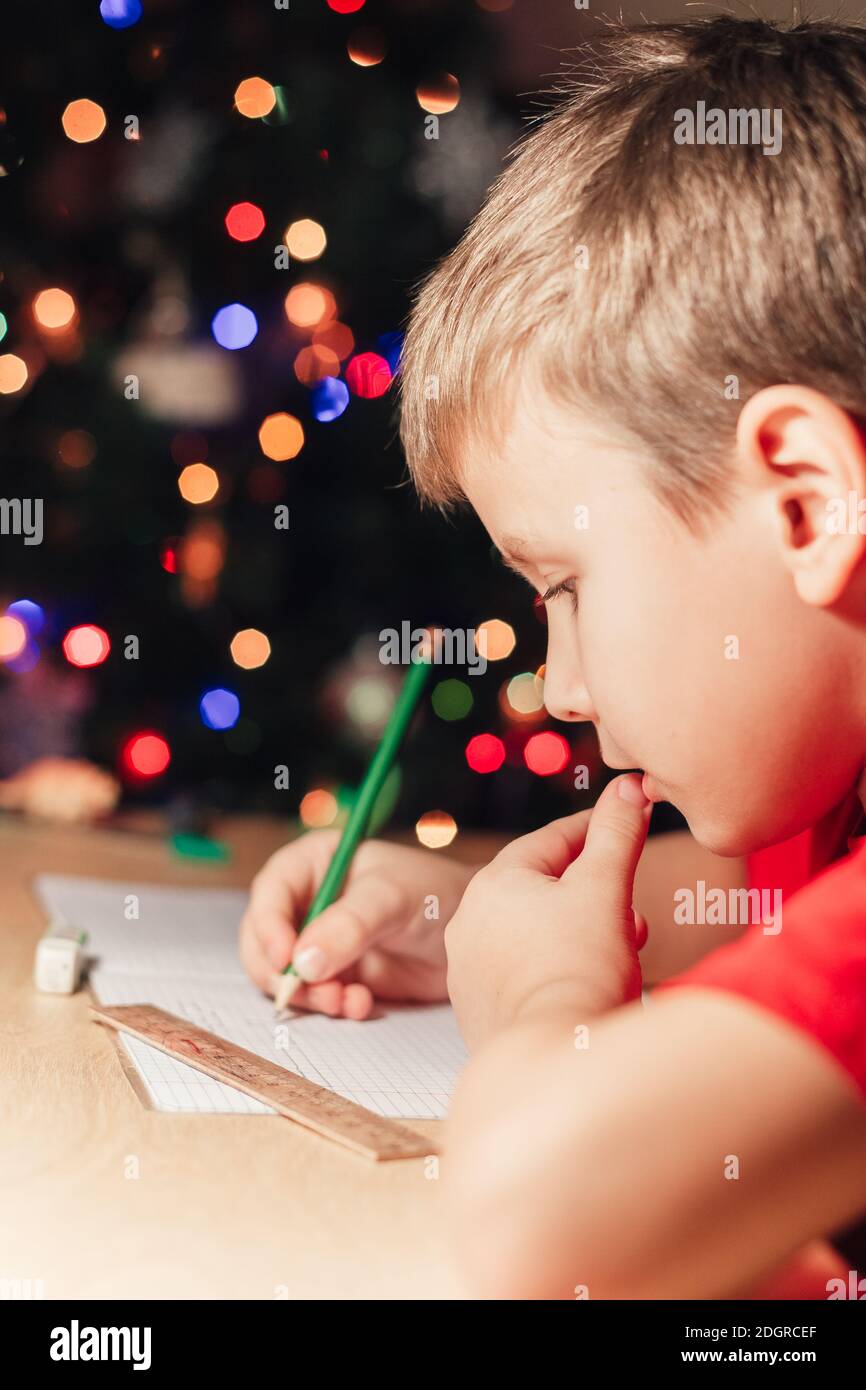 7 Jahre alter Junge, der am Schreibtisch sitzt und Hausaufgaben macht. Kind schreibt Notizen im Notizbuch, schwierige Hausaufgaben, Kind konzentriert sich auf Beispiele. Geschmückter Weihnachtsbaum im Hintergrund Stockfoto