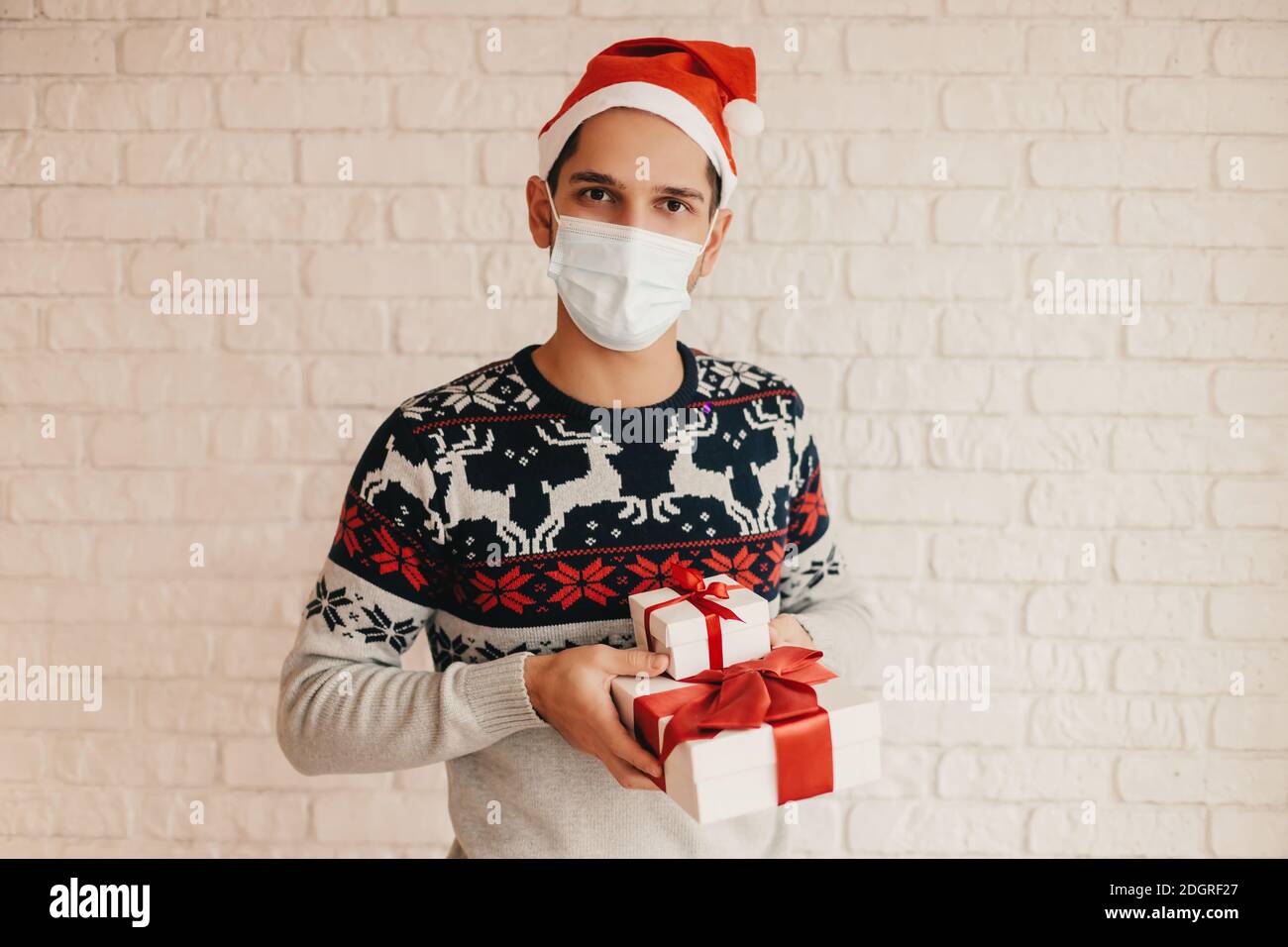 Glücklicher Mann in medizinischen Gesichtsmaske, Weihnachts-Pullover halten Geschenkboxen mit roter Schleife in den Händen. Junge festliche Mann in Santa Hut, Gesichtsmaske mit Neujahr prese Stockfoto