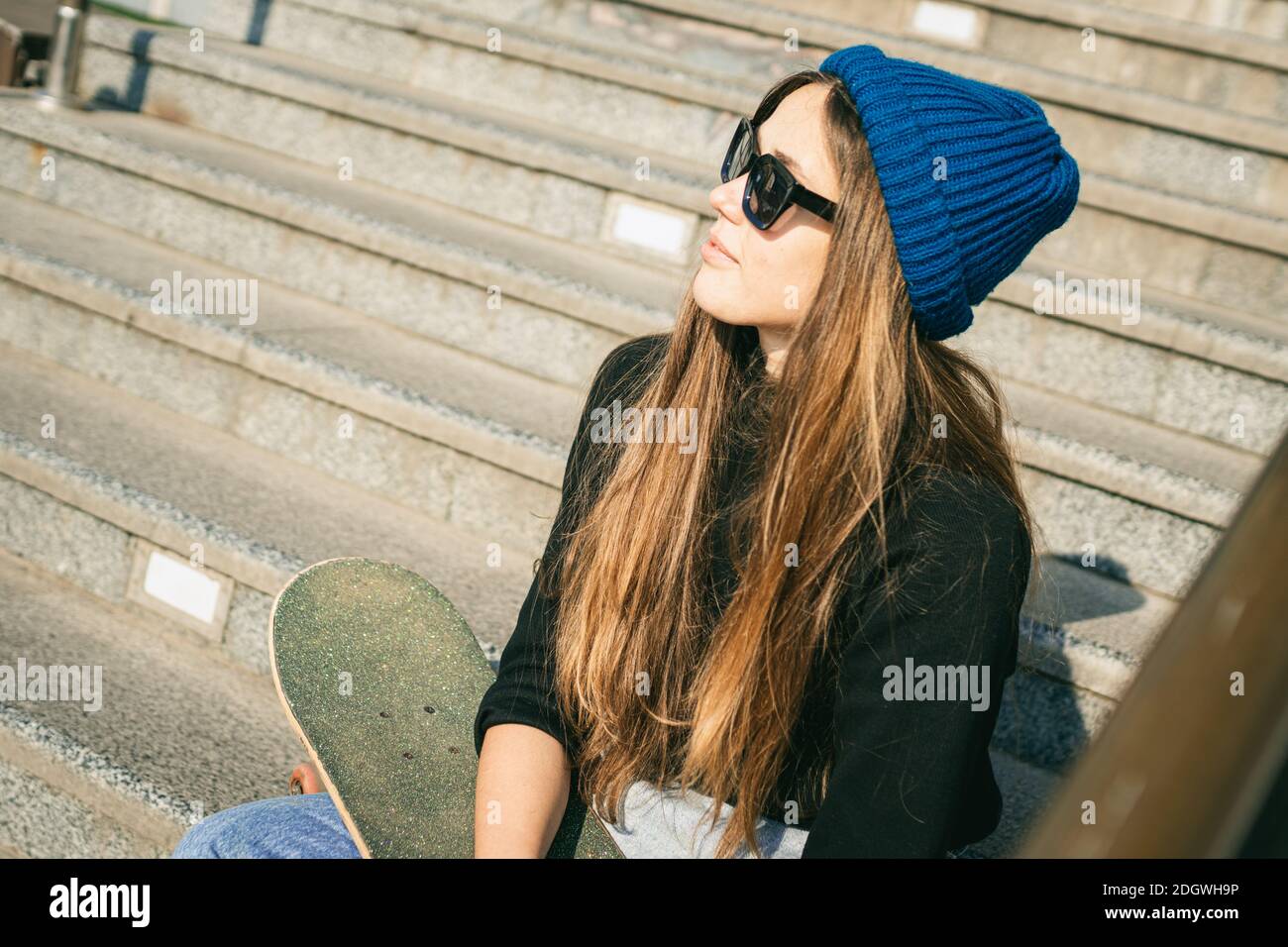 Stilvoll gekleidete Frau in blauem Jeansjumpsuit posiert mit Skateboard. Straßenfoto. Portrait von Mädchen halten Skateboard. Lebst Stockfoto