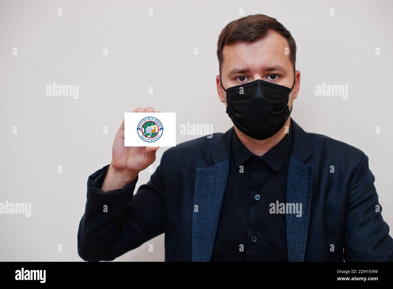 Amerikanischer Mann tragen schwarz formell und schützen Gesichtsmaske, halten Elk Grove Flagge Karte isoliert auf weißem Hintergrund. USA Cities Coronavirus Covid Konzept. Stockfoto