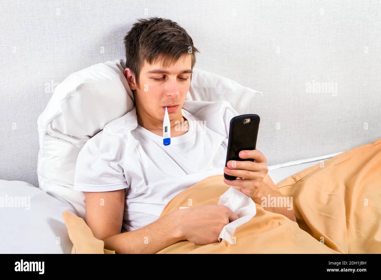 Kranker junger Mann mit einem Telefon und einem Thermometer in Das Bett im Haus Stockfoto