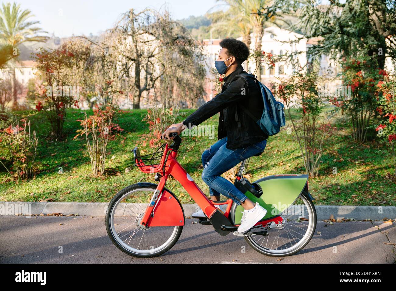 Junger Mann ein marokkanischer Student auf einem gemeinsamen E-Bike in einem schönen Park mit vielen Bäumen bei Sonnenuntergang, trägt eine Gesichtsmaske für die Coronavirus pand Stockfoto