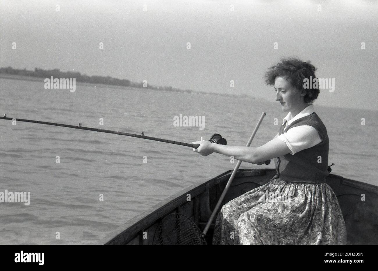 1950er Jahre, historisch, draußen auf See, in einem Holzboot, eine Dame mit einem Rock mit Blumenmuster, einer Oberteil- und Kurzarm-Wolljacke, Angeln, sitzend mit einer Angelrute, mit einem Fischernetz neben ihr, Deal, Kent, England. Stockfoto