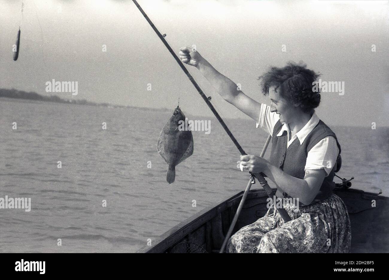 1950er Jahre, historisch, draußen auf See, in einem Holzboot, eine Dame mit gemustertem Rock, Oberteil und Kurzarm-Wolljacke, Angeln, sitzen mit einer Angelrute und halten ihren Fang, einen flachen Fisch, einen Flunder, Deal, Kent, England. Stockfoto