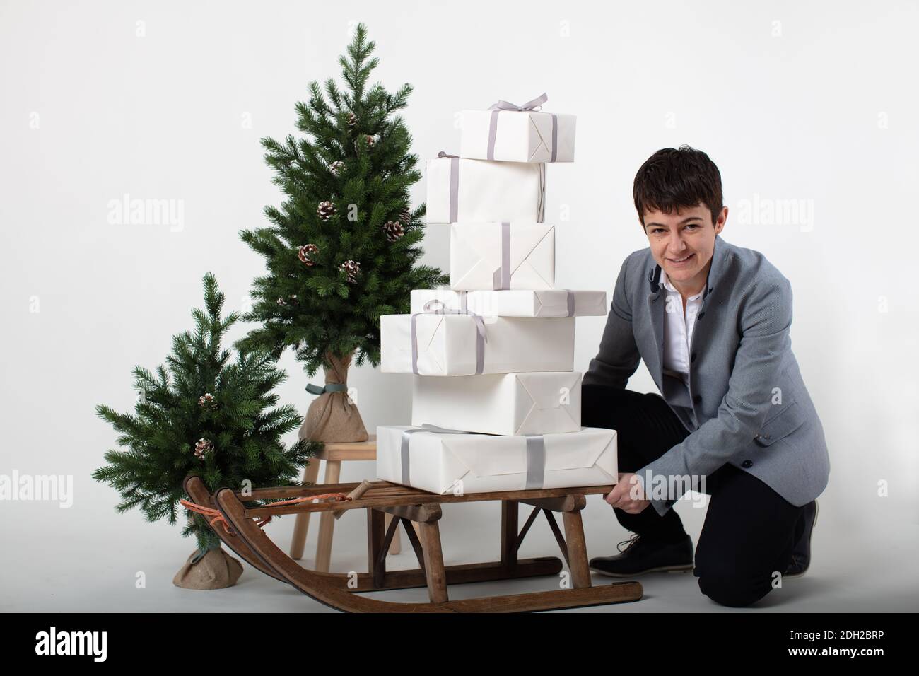 Weihnachts-themed horizontale Business-Porträt eines smart casual gekleidet weibliche Executive schieben einen Schlitten mit weißen und silberumhüllten Geschenkboxen auf einem grauen Hintergrund geladen. Stockfoto