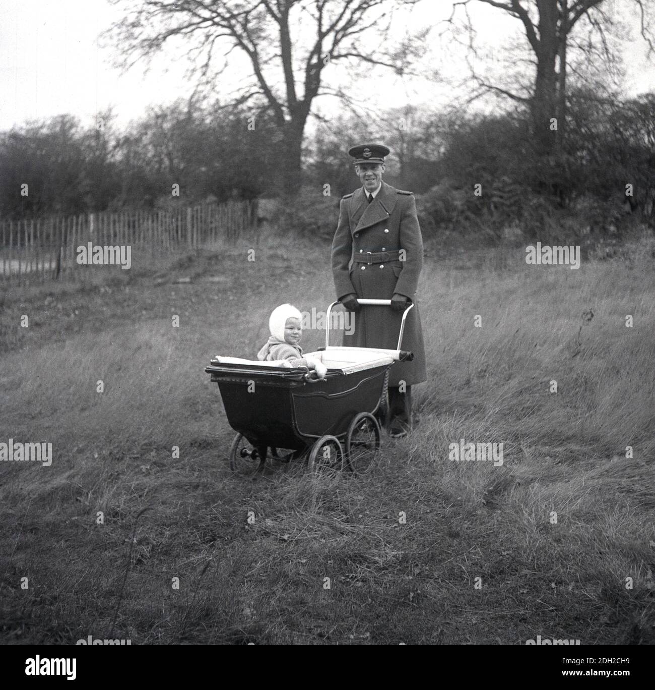 1950er Jahre, historisch, ein Armeeoffizier, in einem langen Militärmantel und Hut auf einem Feld aus langem Gras, der den Griff eines Kutschwagen der Ära hält, in dem sein Kleinkind oben sitzt, England, Großbritannien. Stockfoto