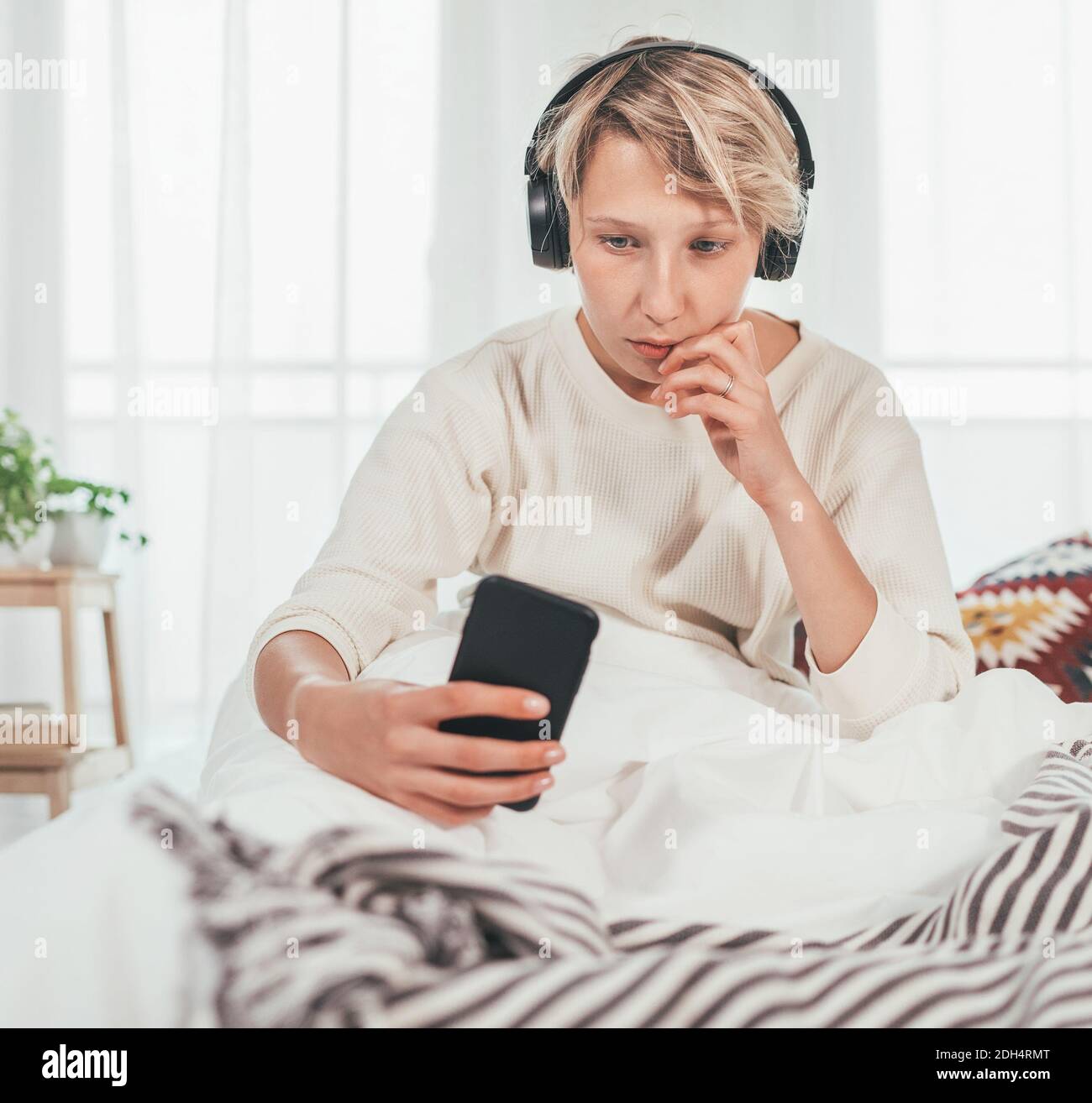 Junge Frau sitzt auf einem gemütlichen Bett gekleidet Schlafanzug im Internet surfen und Musik hören mit kabellosen Kopfhörern. Musik spielt moderne Technologie ein Stockfoto