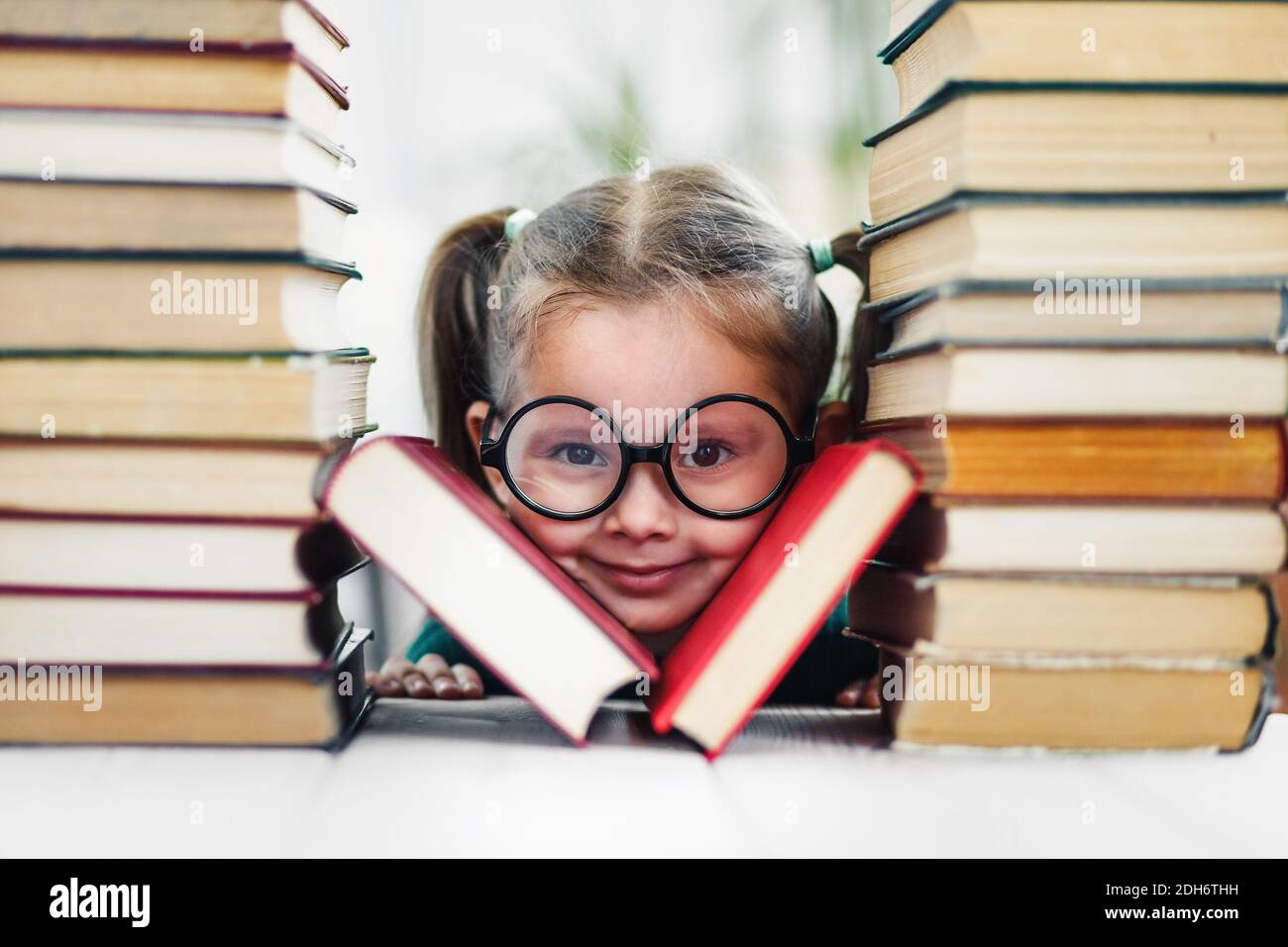 Vorschulalter lächelndes Mädchen mit Grübchen in großen lustigen Gläsern zwischen einem Stapel von Büchern. Stockfoto