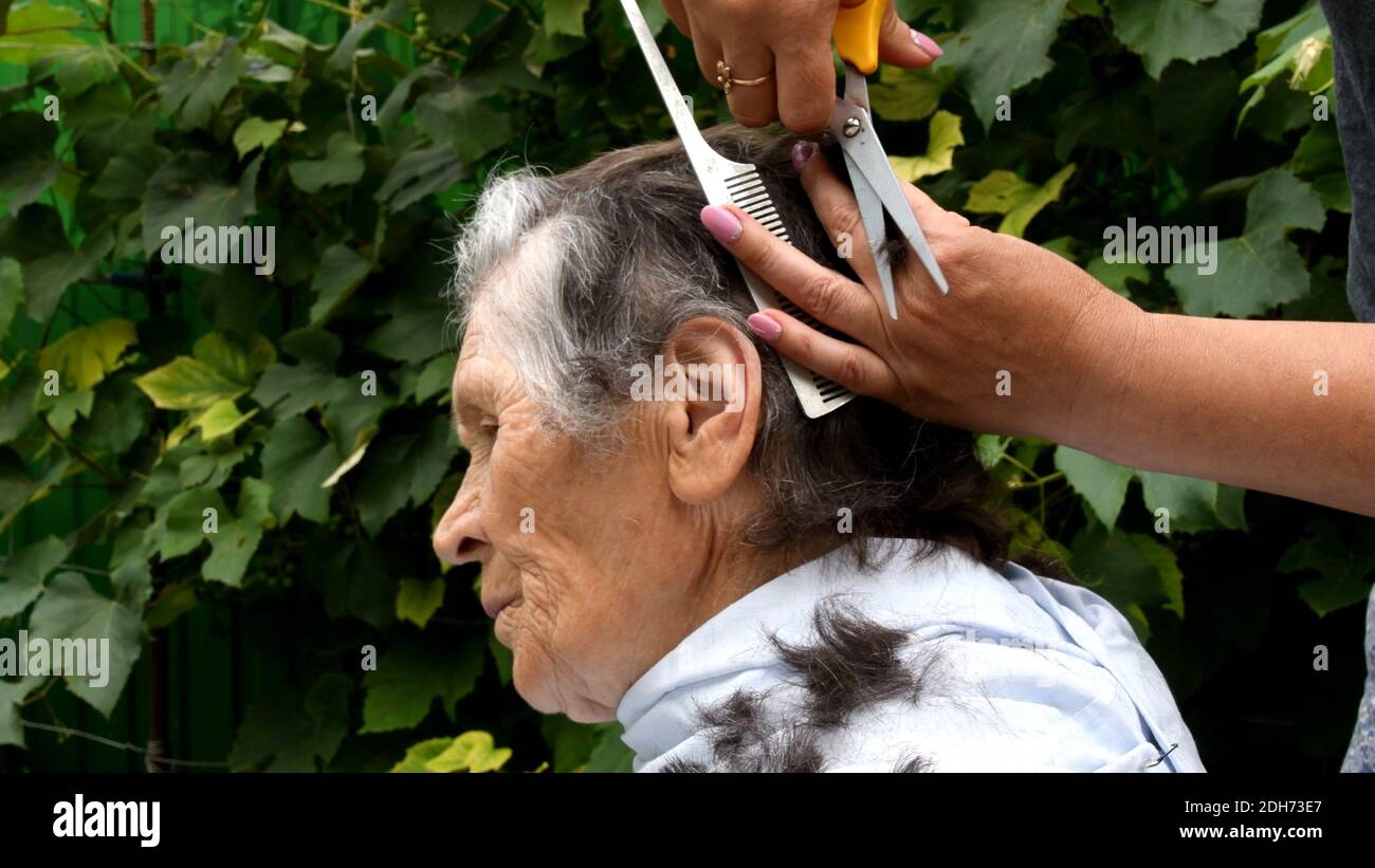 Weibliche Hände schneiden Haare von älteren Frau zu Hause im Hinterhof im Freien. Frau Hände hält Kamm und Schere, um Haare von 80 Jahre alte Frau zu schneiden Stockfoto