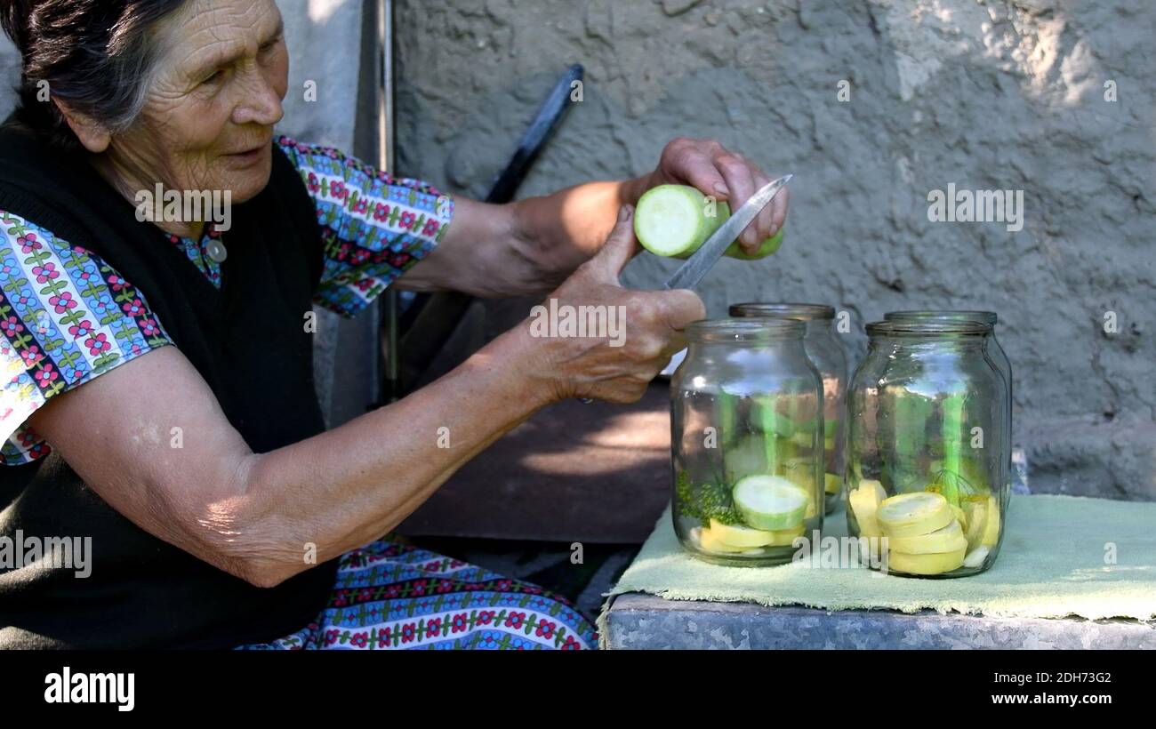 Ältere asiatische Frau lächelt und schneidet frische Zucchini mit Küchenmesser. Hausgemachtes Einmachtes. Oma bereitet Gemüse als Konservierung in Gläsern vor Stockfoto