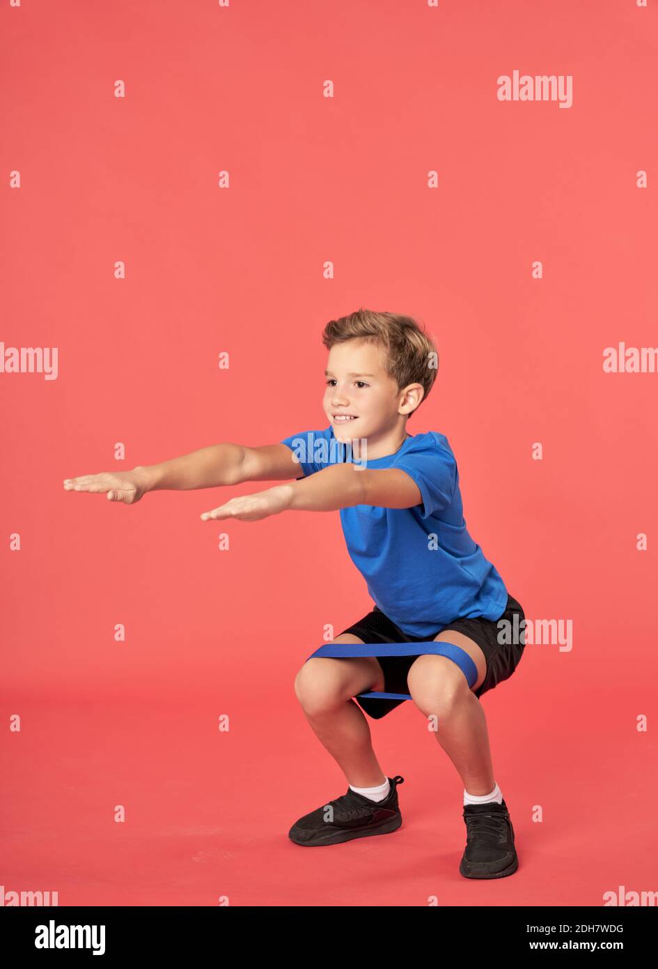 Nettes männliches Kind schaut weg und lächelt beim Hocken Mit Widerstandsband vor rotem Hintergrund Stockfoto