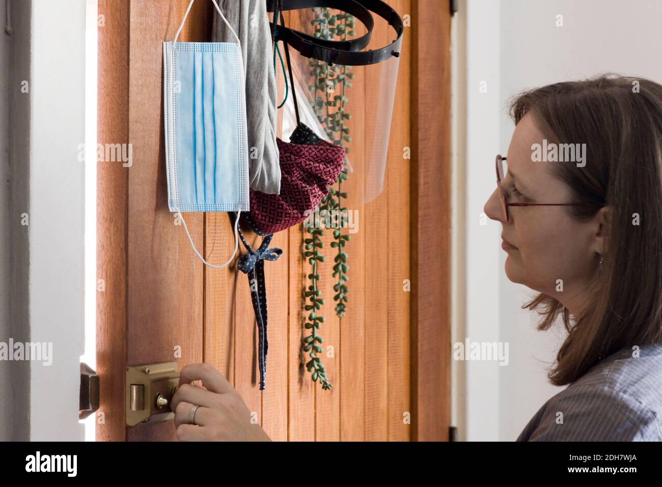 Eine Frau schließt ihre Haustür, während sie sich eine Auswahl an Schutzausrüstung einschließlich einer Einweg-Gesichtsmaske, Stoffmaske und einem Gesichtsschutz ansieht. Stockfoto