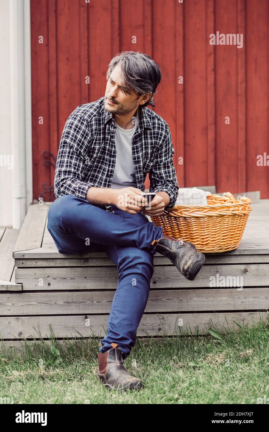 Nachdenklicher Bauer mit Korbkorb, der das Mobiltelefon hält, während er sitzt Auf der Veranda im Hof Stockfoto