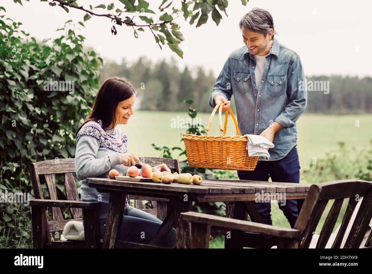 Glücklicher Mann, der Korb auf den Tisch legt, während Frau Äpfel schneidet Auf Bio-Bauernhof Stockfoto