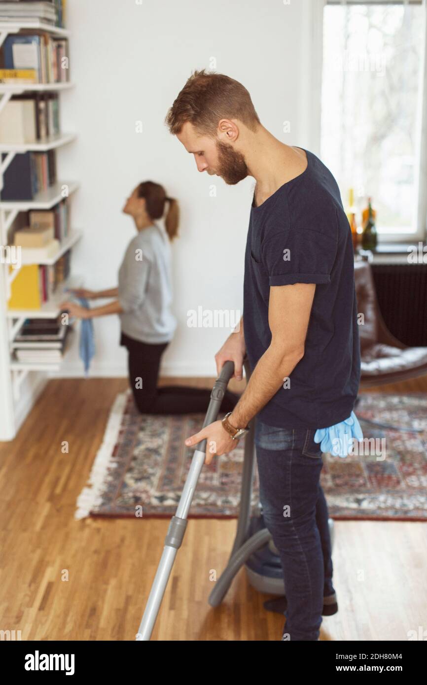 Seitenansicht des Mannes, der den Boden saugt, während die Frau die Regale putzt Im Hintergrund zu Hause Stockfoto