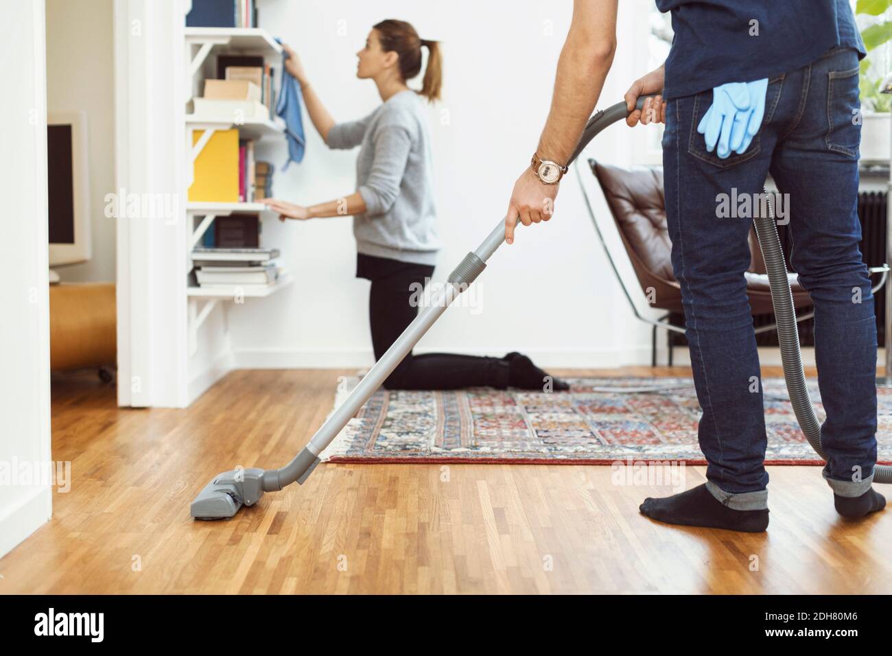Niedriger Abschnitt des Mannes, der Boden saugt, während Frau Regale putzt Im Hintergrund zu Hause Stockfoto