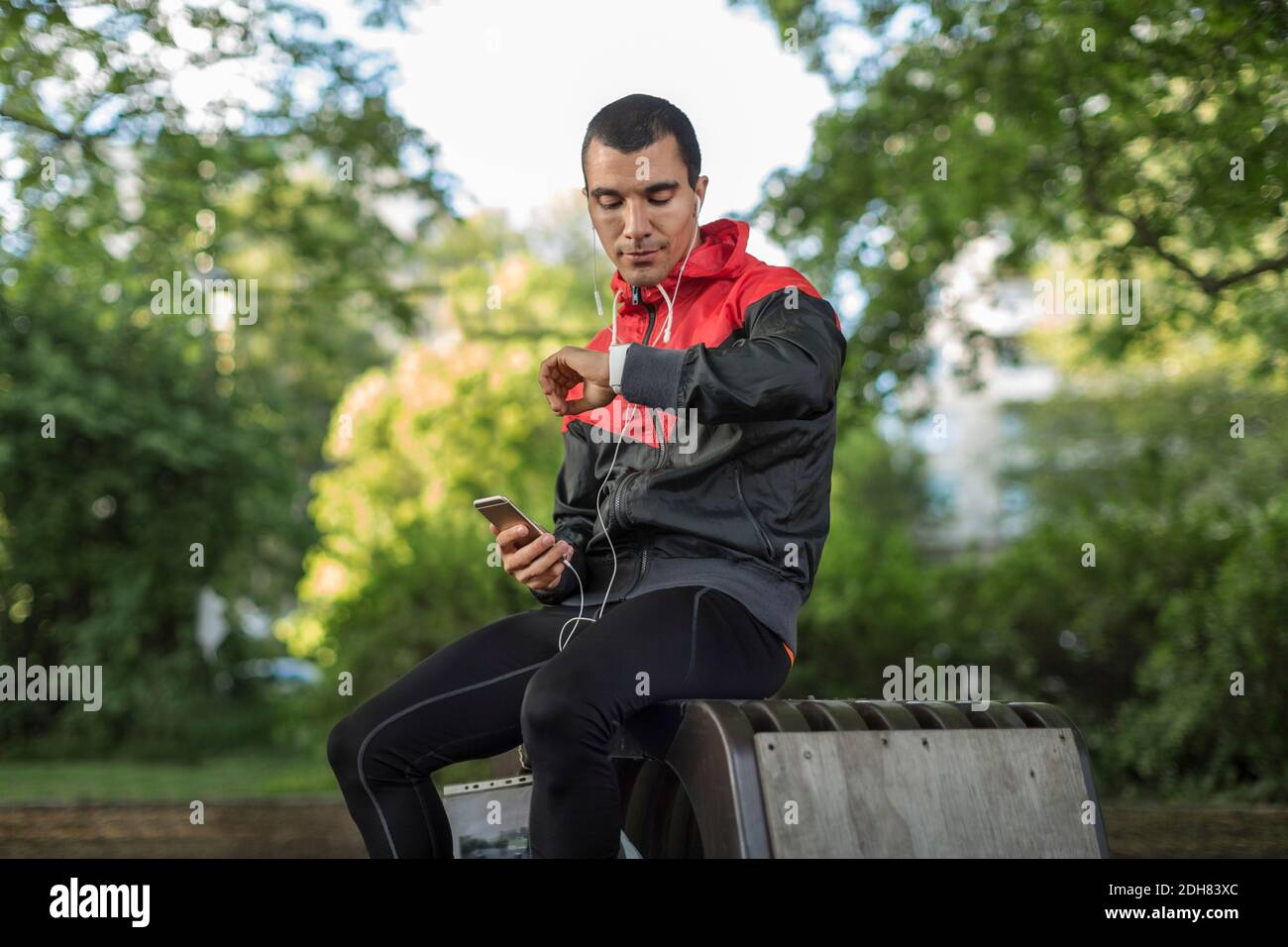 Mann, der auf der Bank sitzt und das Telefon hält und auf die Armbanduhr schaut Stockfoto
