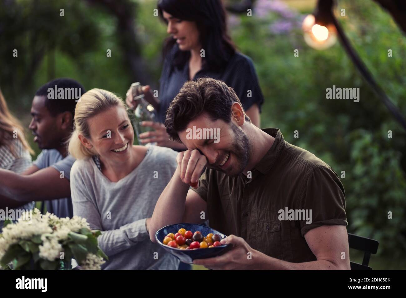 Glückliche Frau, die den Mann ansieht, der die Augen reibt, während sie Tomate hält Schüssel bei der Dinner-Party im Hof Stockfoto