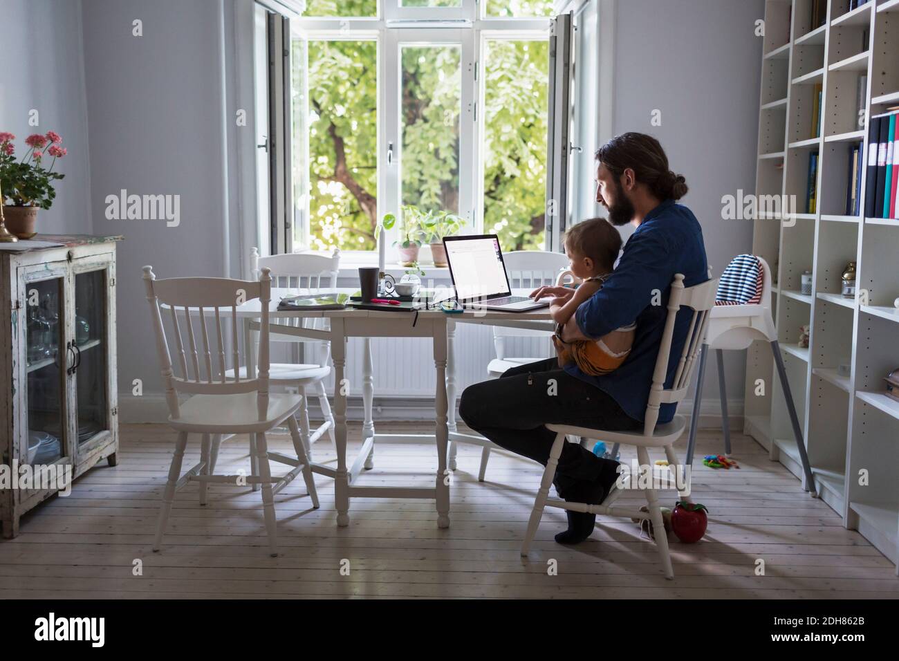 Volle Länge des Vaters mit Laptop-Computer, während die Pflege Von Baby zu Hause Stockfoto