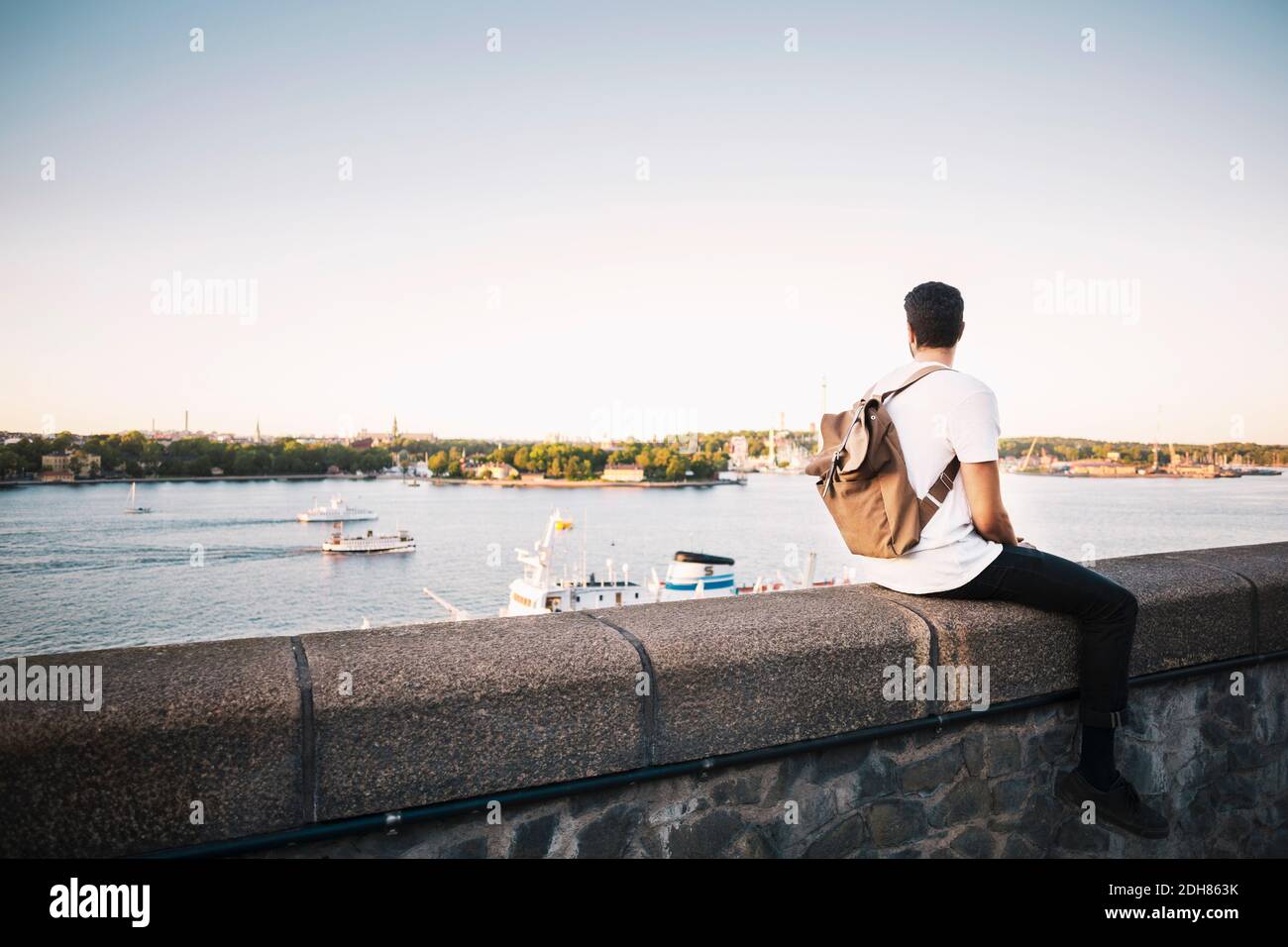 Männlicher Tourist, der die Aussicht ansah, während er auf einer Stützmauer saß Von Brücke gegen klaren Himmel Stockfoto