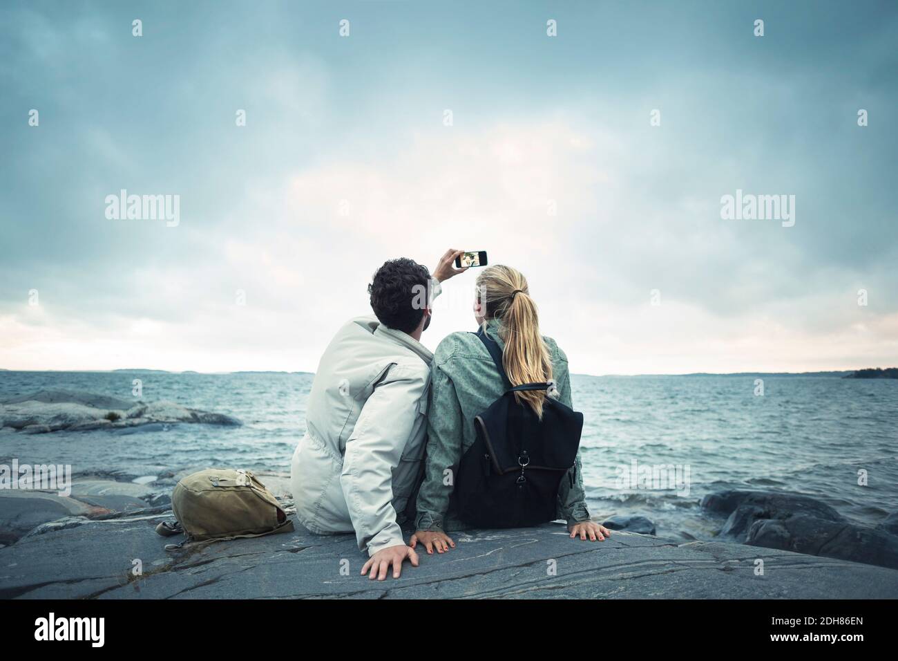 Ein Paar nimmt Selfie, während es auf Felsen am Meer sitzt Stockfoto