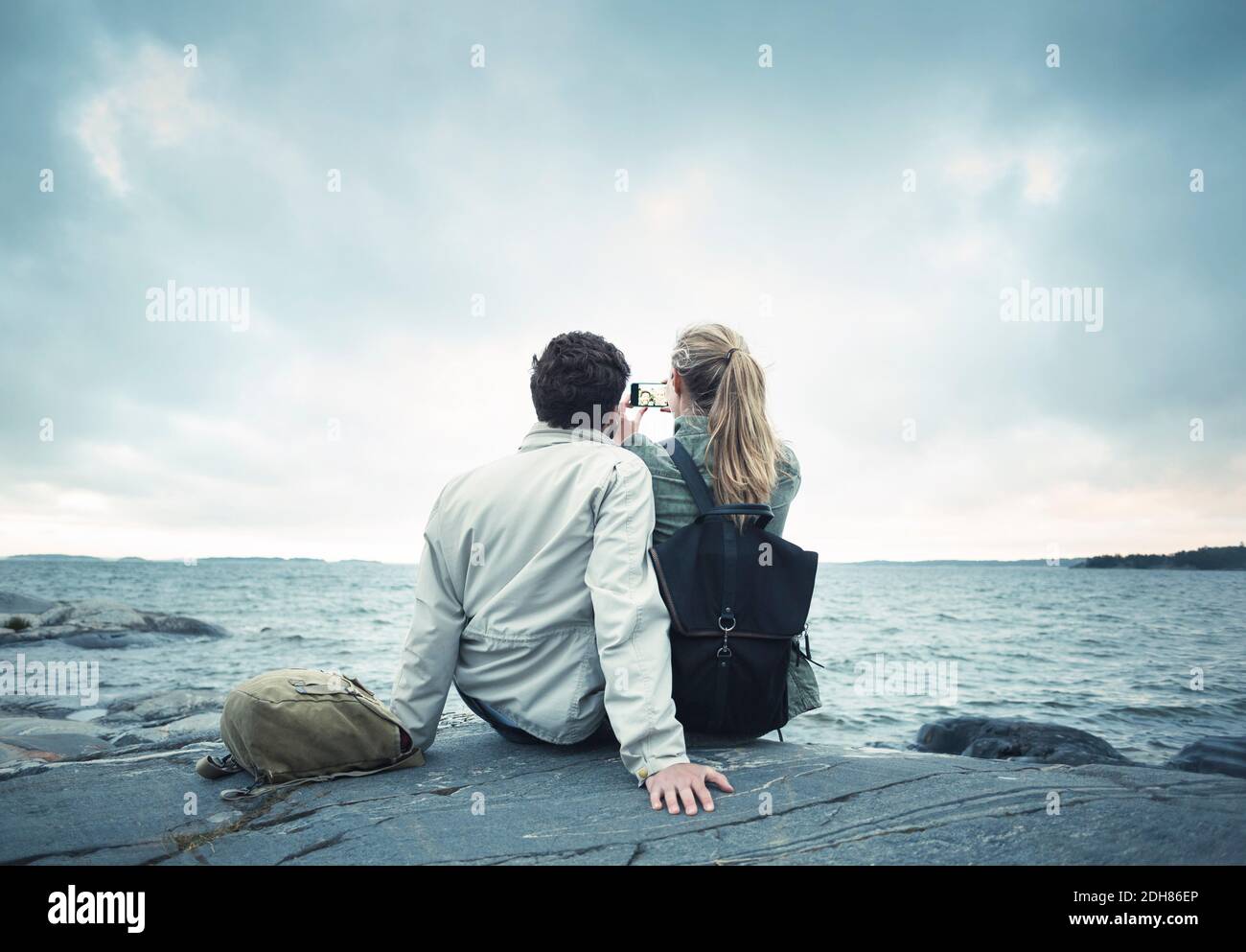 Rückansicht des Paares, das Selfie beim Sitzen auf Felsen nimmt Auf dem Seeweg Stockfoto