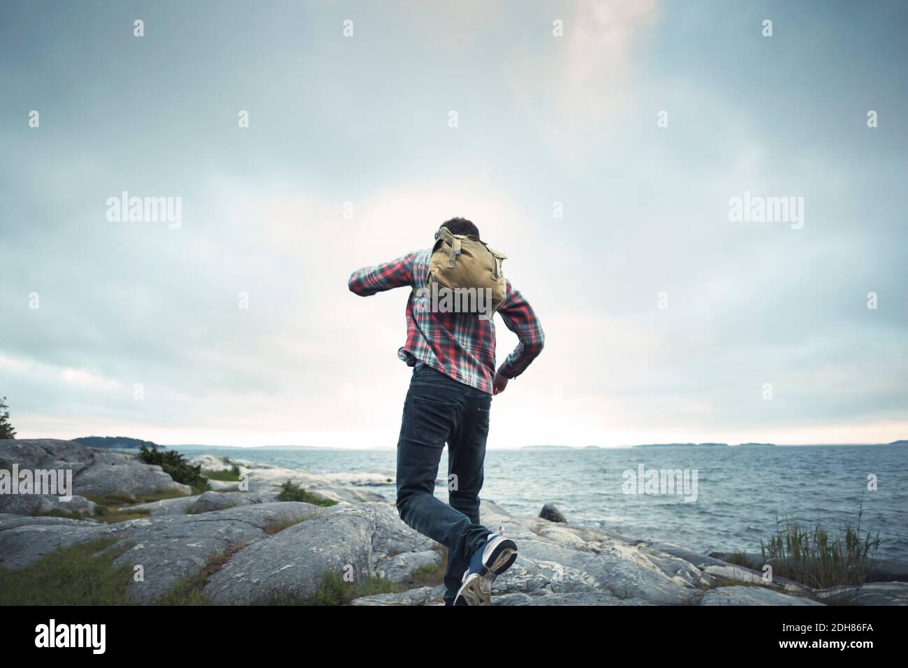 Rückansicht des wonderlust-Mannes, der auf Felsen am Meer springt Es weht schwacher bewölktes Wetter Stockfoto