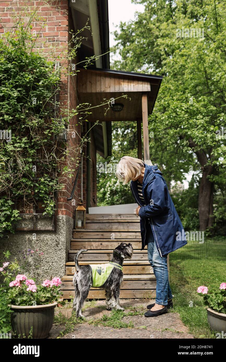 Seitenansicht einer älteren Frau, die im Stehen eine Jacke trägt Hund vor dem Haus Stockfoto