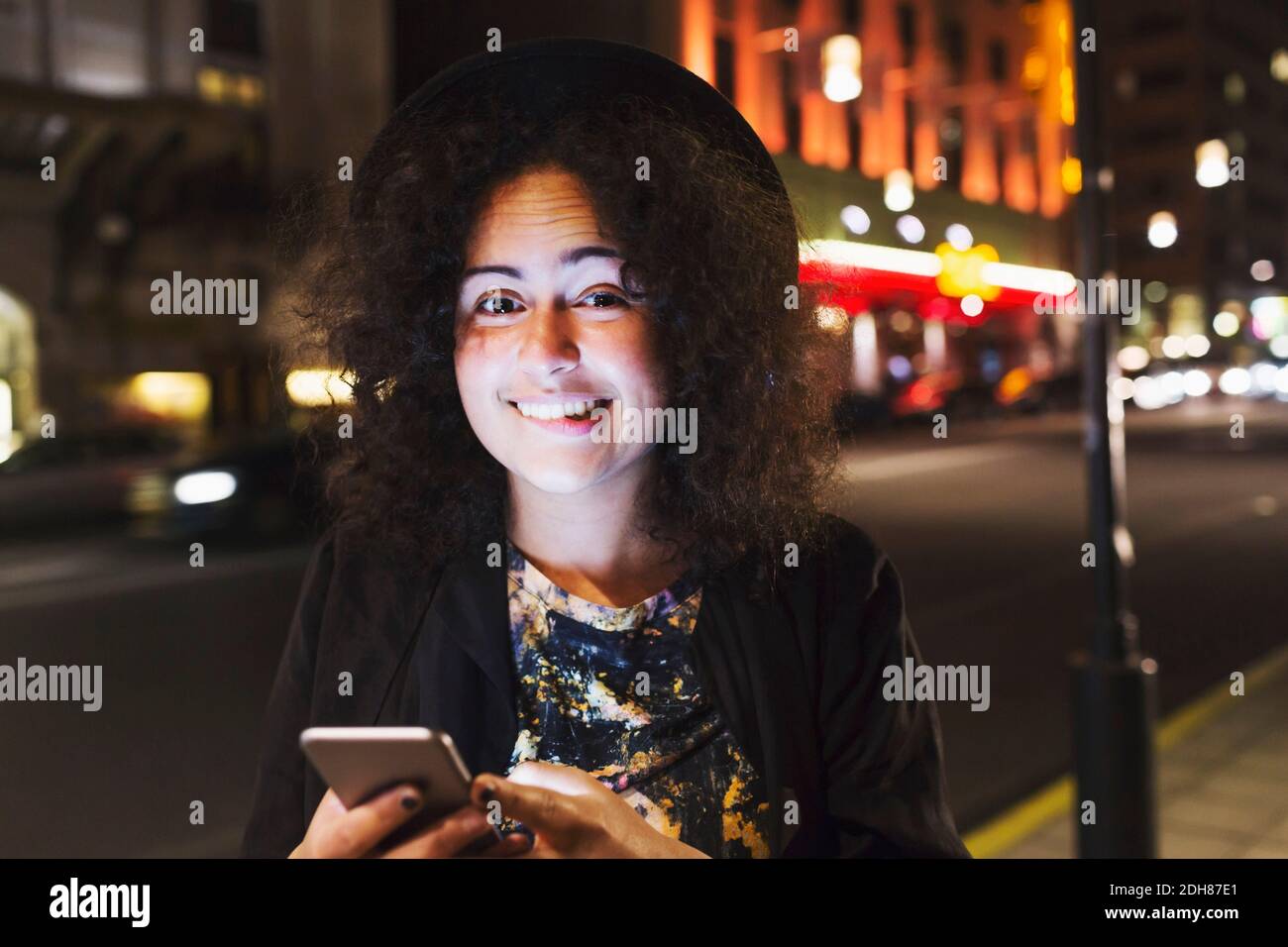 Porträt einer lächelnden Frau beißt Lippe, während das Smartphone halten Nachts in der Stadt Stockfoto