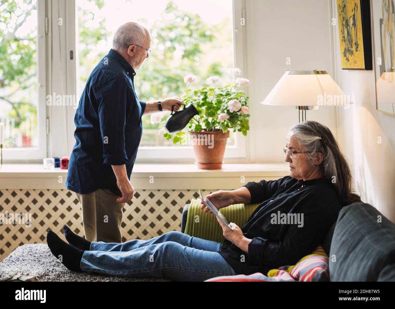 Volle Länge der älteren Frau mit digitalen Tablet, während Mann Bewässerung Blume Pflanze zu Hause Stockfoto