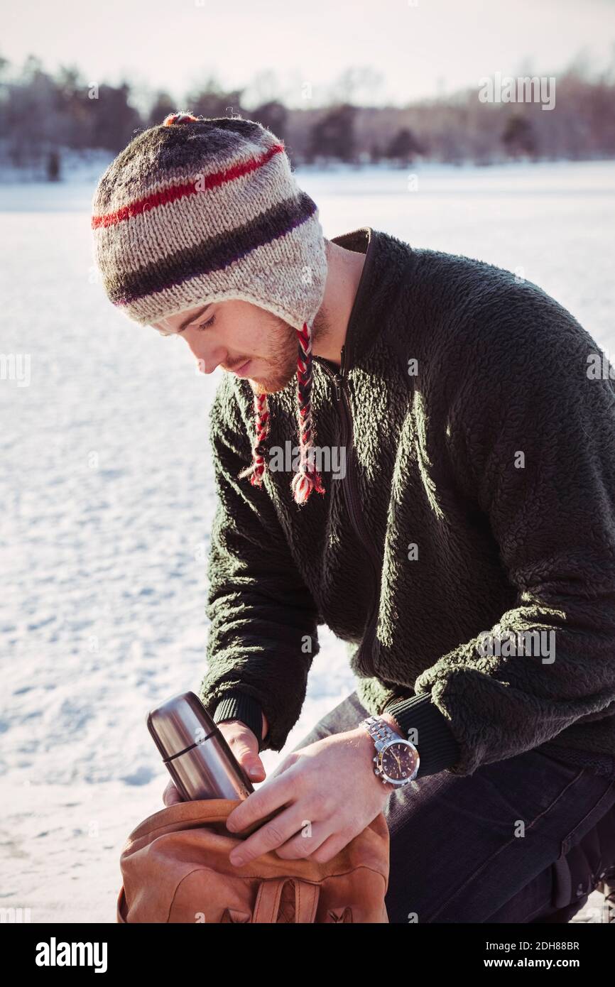 Junger Mann, der Thermoskannen in den Beutel legt, während er auf Schnee kniet Abgedecktes Feld Stockfoto