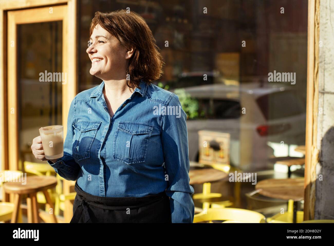 Glückliche Besitzerin hält Kaffeetasse, während sie gegen das Café steht Stockfoto