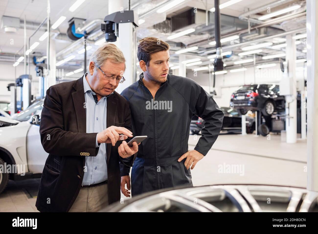 Kunde, der ein Mobiltelefon verwendet, während er mit einem Mechaniker im Auto steht Reparaturwerkstatt Stockfoto