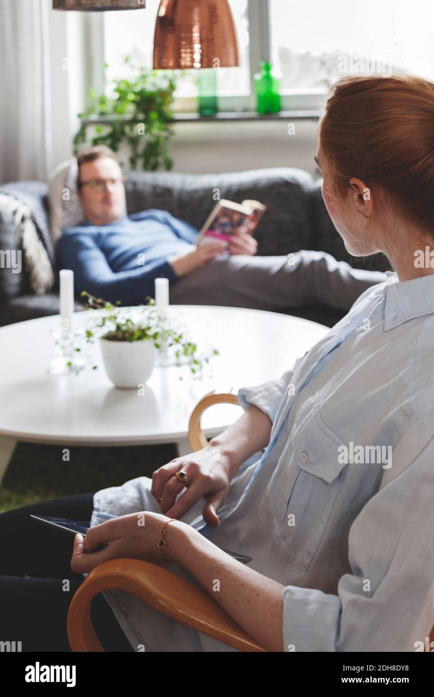 Frau mit digitalen Tablet beim Blick auf Mann Buch lesen Im Wohnzimmer Stockfoto
